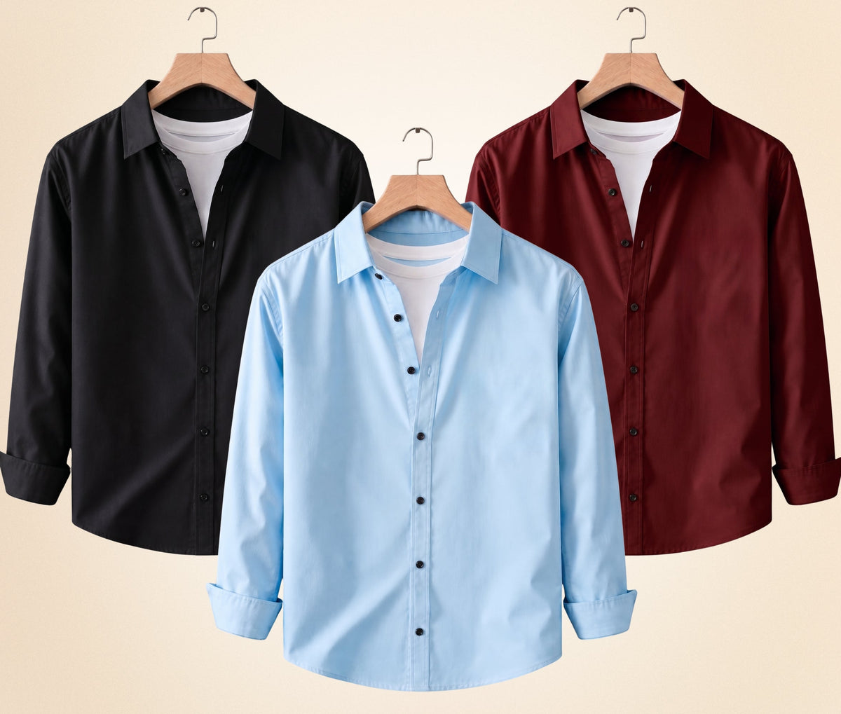 Three long-sleeve button-up shirts on wooden hangers against a light beige background: a black shirt on the left, a light blue shirt in the center, and a burgundy shirt on the right. Each shirt is layered over a white undershirt.